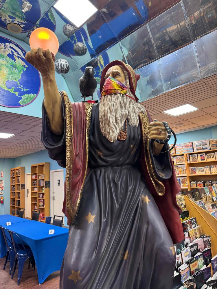 A statue of an old wizard with a long white beard and elaborate robe and hood rising a crystal ball above him and looking into it. The crystal ball has shifting color lights.