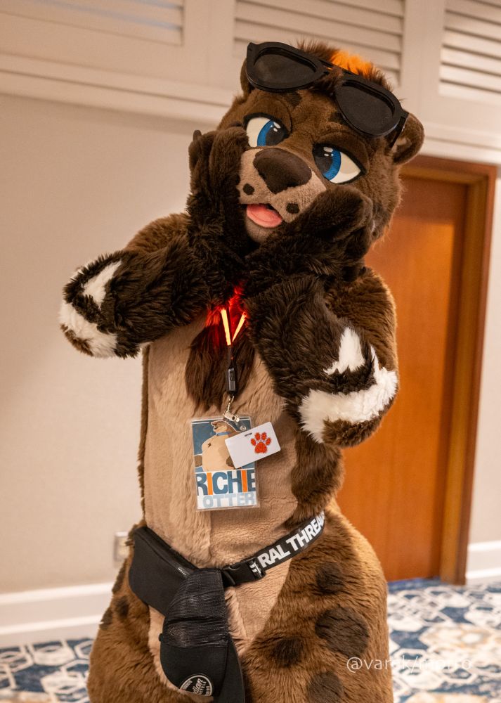 Hotel interior. Photo of Richie (brown and tan otter fursuiter) standing, with a surprised expression. 
