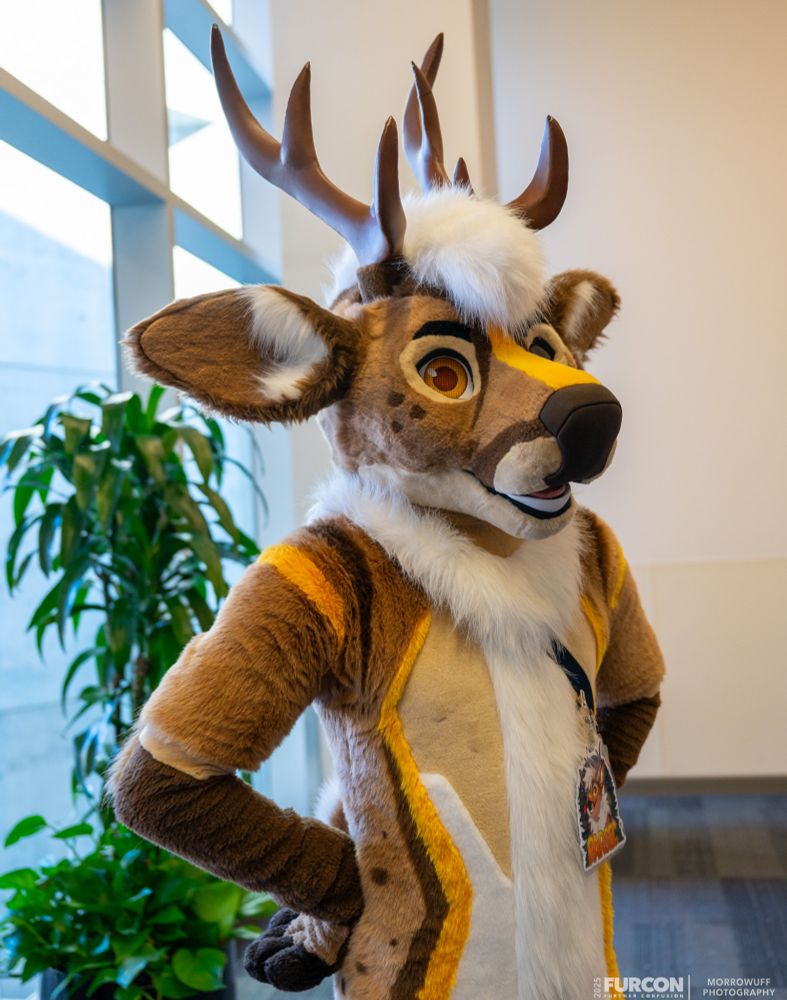 San Jose convention center, inside. Photo of Parsec (brown, white, and tan deer fursuiter with dark brown and yellow markings and antlers), hands on hips, striking a regal pose