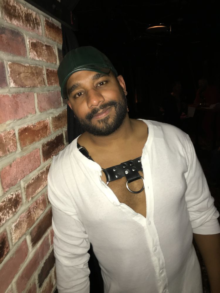 An older photo of me in Philly in 2016. I’m in a bar, photographed leaning against a brick wall with a dark background. I’m wearing a white union suit with a black leather harness underneath, a hunter green leather cap, and an obnoxious bearface smirk 🤣