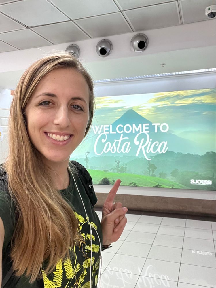 Isa smiling at the camera and pointing to a “welcome to Costa Rica” tourism sign that is in the background. 