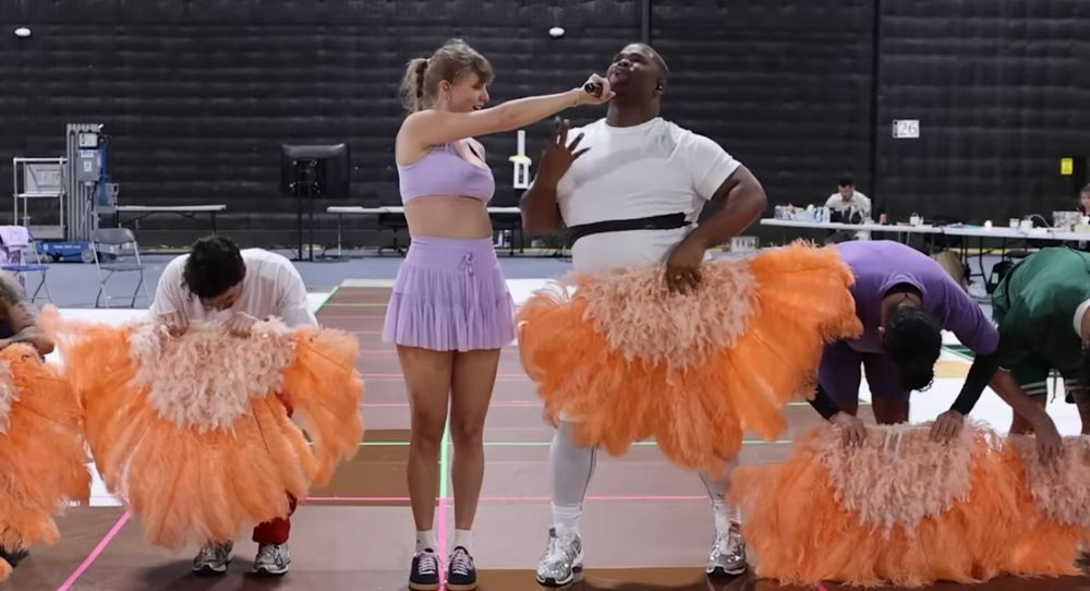 Still from “I can do it with a broken heart” music video. Taylor swift (wearing lavender skirt and top) holds the microphone up to dancer Cam. Dancers are bent over, holding orange feathers. 