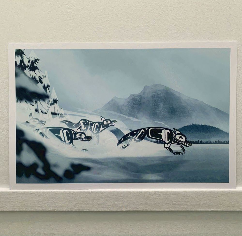 A picture of an art print tacked against a white wall. The print depicts a pack of black and white formline wolves charging across a frozen lake while mist swirls around them, with foggy mountains and hills in the distance.