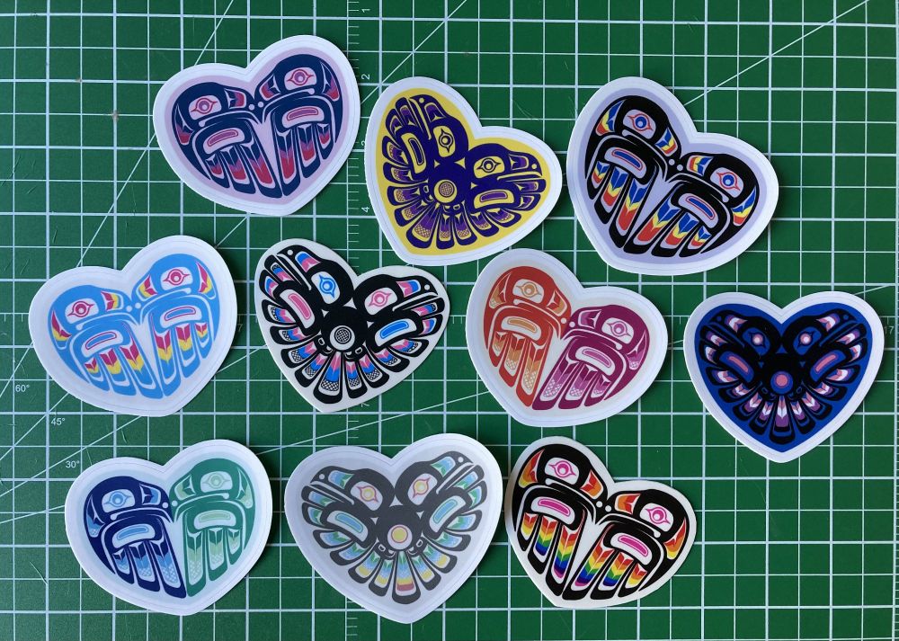 A picture of a bunch of formline stickers- both have designs of mirrored formline ravens, one design looking inward and the other outward. They are coloured in various pride flag palettes.