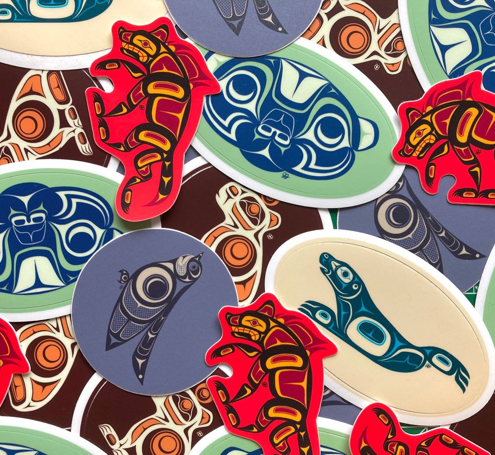 A picture of an assortment of animal stickers spread out and laying on top of each other. All of them are formline designs, and show the following: a blue and cream-coloured chickadee, a yellow and blue-teal harbour seal, a maroon and orange capybara, a light green and dark blue otter head, and a bright red and dark red hyena.