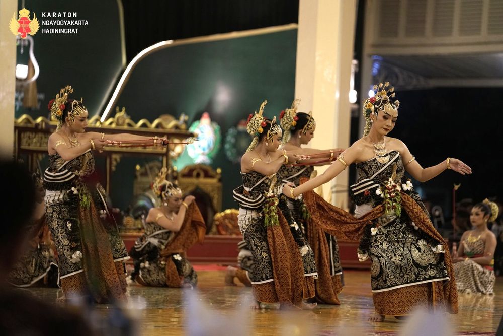 A photo of Yogyakarta court dancers performing Bedhaya Mintaraga. Several women in gold crowns and jewelry, black embroidered dresses, and batik skirts and sashes danced with bows and arrows. Behind them is a gamelan set.