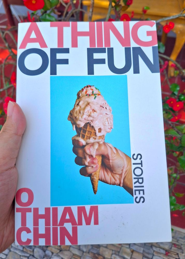A photo of my hand holding A Thing of Fun: Stories book by O Thiam Chin. The book cover is white with a photograph of a hand holding a melting pink funfetti ice cream cone with light blue background. 
