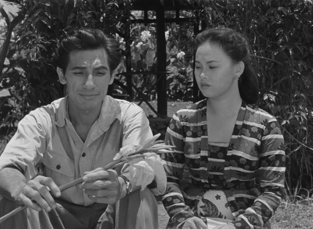 A clip from Lewat Djam Malam, depicting lovers Iskandar and Norma sitting together. Iskandar holds a flower while Norma looks on, their expressions are clouded with gloom despite Iskandar's smile.