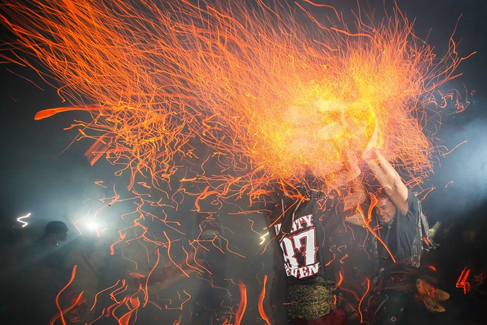 Two men in black shirts and decorated sashes around their hips clashing burning coconut husks high above their heads in Mesabetan Api ritual.