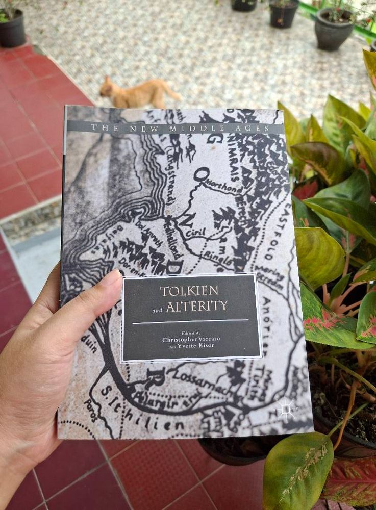 A photo of my hand holding a book titled Tolkien and Alterity, edited by Christopher Vaccaro and Yvette Kisor. The cover art features close up of Tolkien's hand-drawn map of Middle-earth in grey.