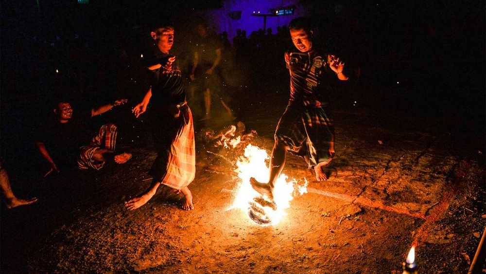 A photo of two Indonesian men playing a fire football, one of them is kicking a flaming ball with a bare foot. Both wear t-shirts and sarongs.