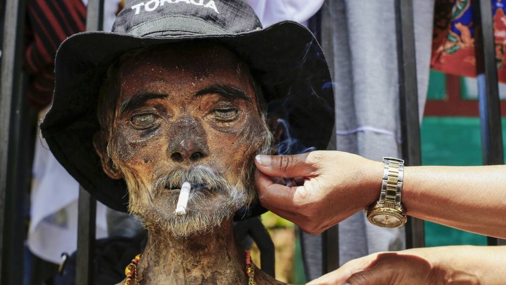 A close up of the face of a man's cadaver with mustache and short beard. He was wearing a black hat that says "Toraja". A pair of hands took care of his facial hair and slipped a cigarette between his lips.