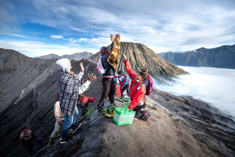A photograph of several Tengger people standing on the edge of the Bromo volcanic crater, surrounded by fog, throwing some fruits and vegetables into the crater.