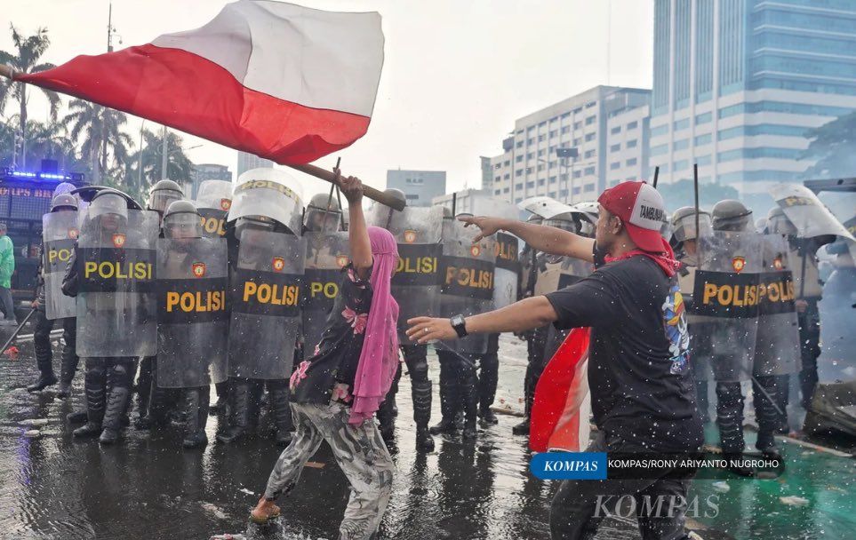A photo of a woman in pink hijab standing her ground against a well-equipped Indonesian riot force. Her thin hands holding an Indonesian flag tied to a bamboo pole, which she waves menacingly. A man, another protester, an Indonesian flag around his neck, tried to stop her.
