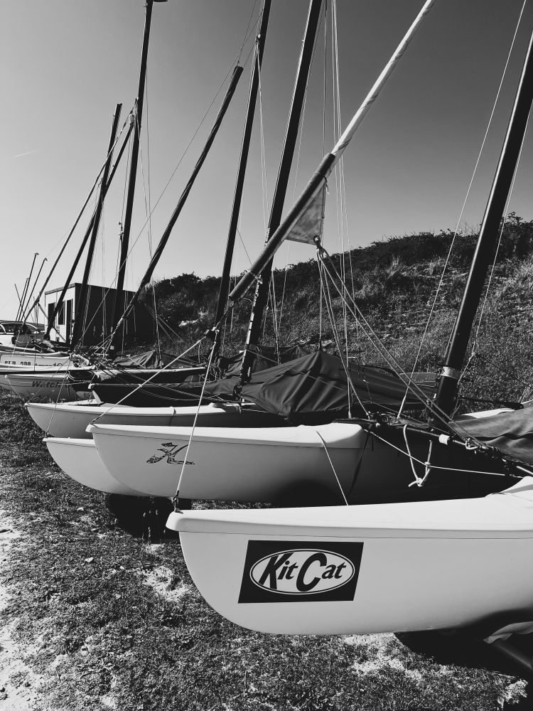Row of little sailing boats in a boatyard. Nearest one is called Kit Cat, design made to look like a popular chocolate bar 