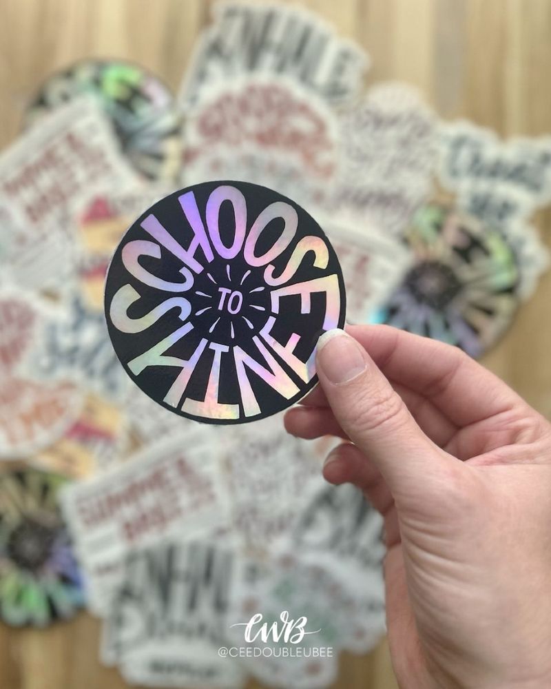 A photo showing an assortment of colorful, hand lettered stickers in the background with my hand in the foreground holding a sticker that reads "Choose to Shine"
