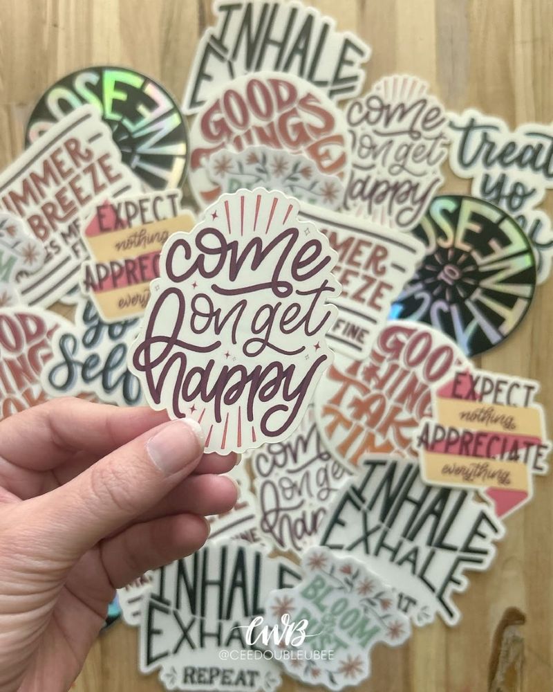A photo showing an assortment of colorful, hand lettered stickers in the background with my hand in the foreground holding a sticker that reads "Come on get happy"