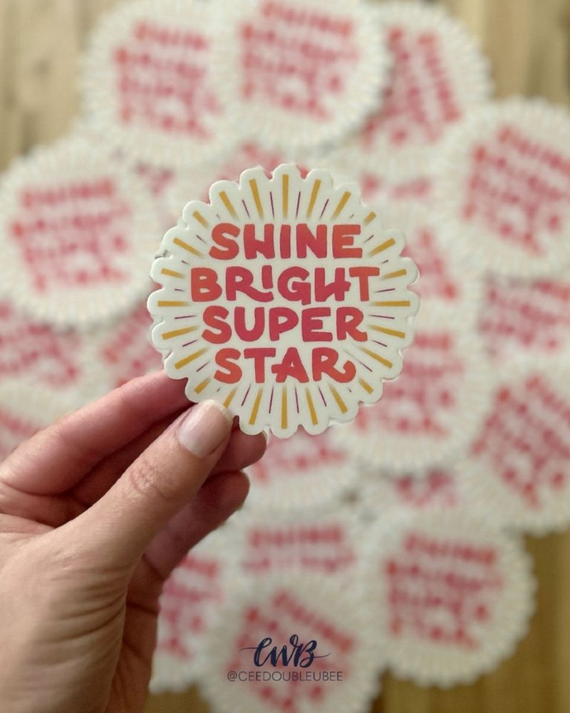 My hand holding the August Sticker: Shine Bright Super Star in bright pink block lettering with an orange-yellow sunburst radiating out from the letters.  More stickers are in the background, blurred behind. 