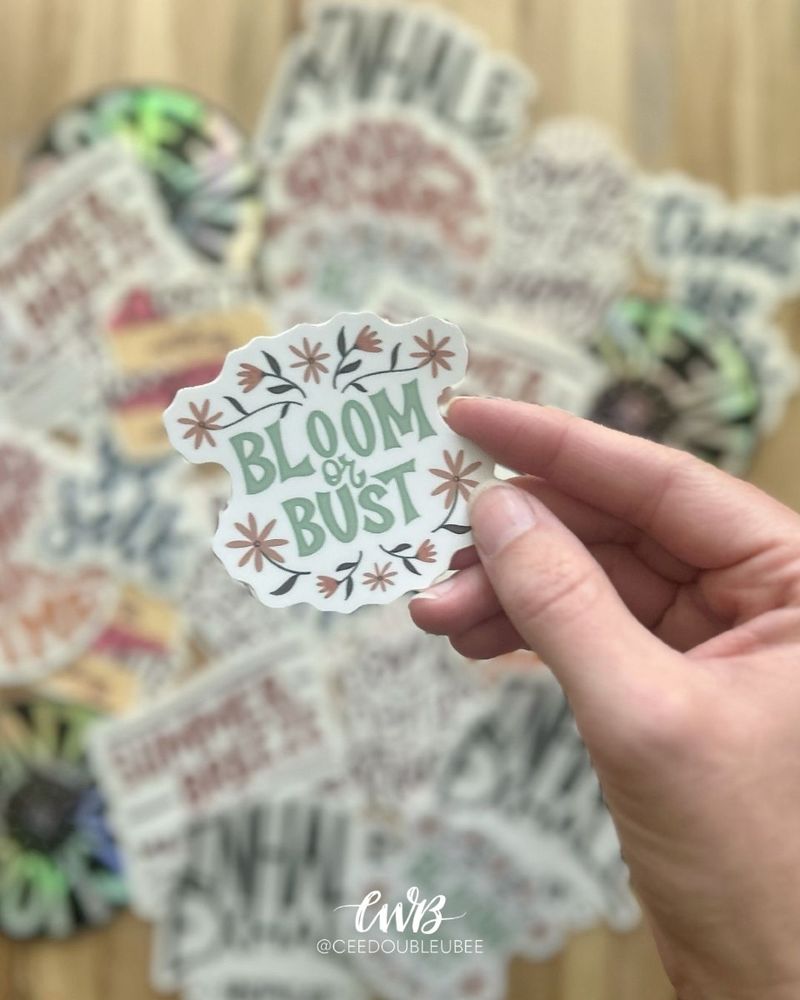 A photo showing an assortment of colorful, hand lettered stickers in the background with my hand in the foreground holding a sticker that reads "Bloom or Bust"