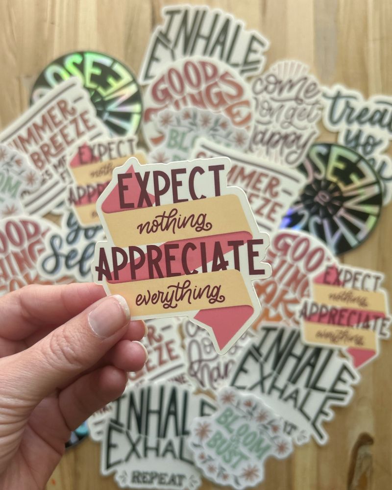 Hand holding a sticker that reads “expect nothing, appreciate everything” with more stickers in the background.