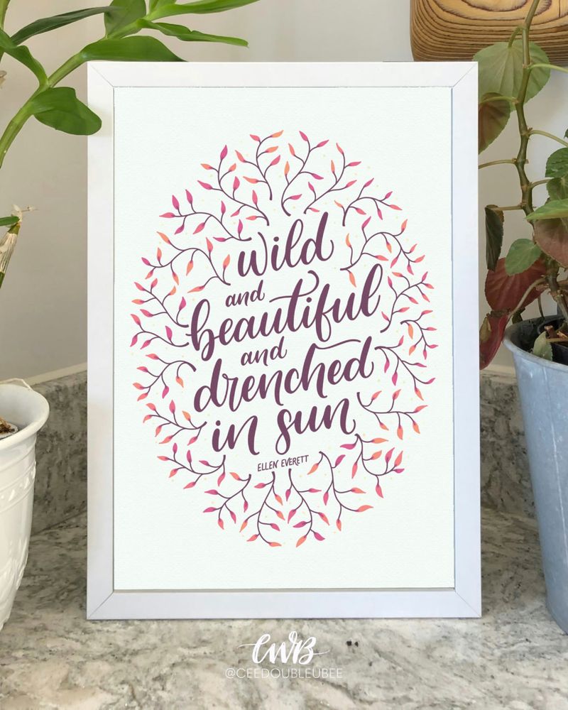A mockup of the “Wild and Beautiful and Drenched in Sun” hand lettered art print, featuring soft pastel summer colors, leafy embellishments, and bold script lettering. The print is shown framed on a countertop with plants on either side.
