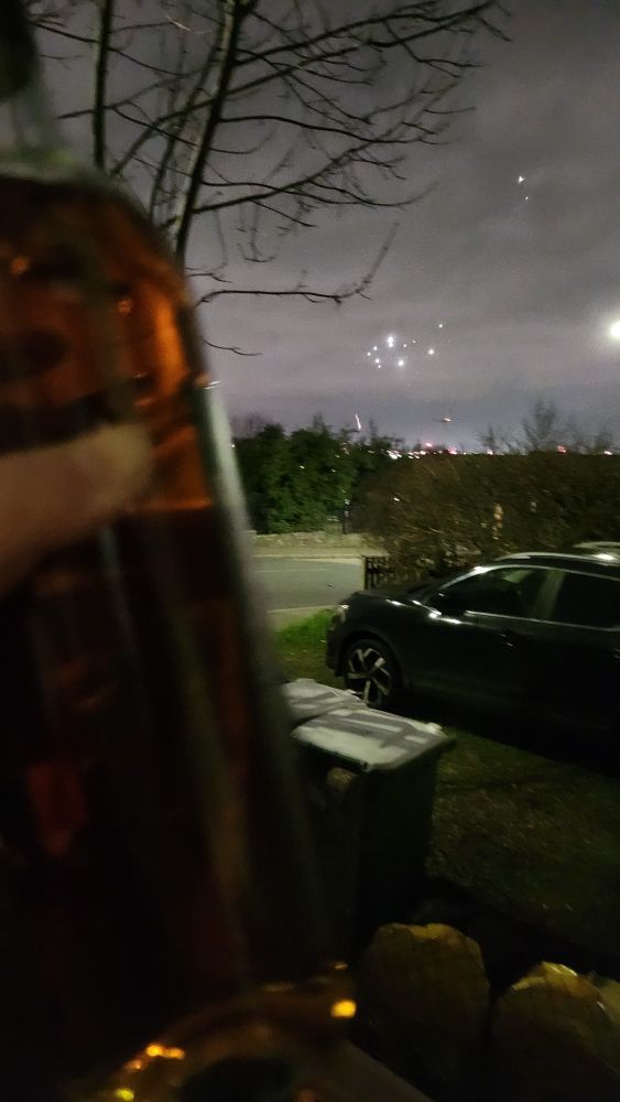 a blurry wine bottle being held up in front of a view of the sky at night, there is a dying firework in view above the trees. A car is visible in the midground