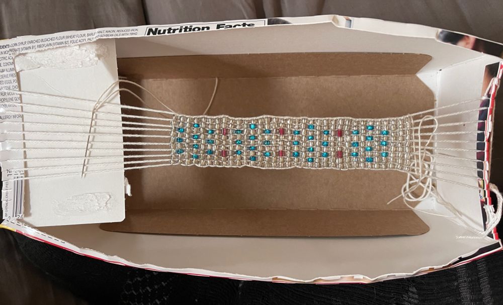 a cardboard box cut out with white thread warped over the gap. beads have been woven in a simple pattern with clear, blue, and red beads. the work is nearly finished 