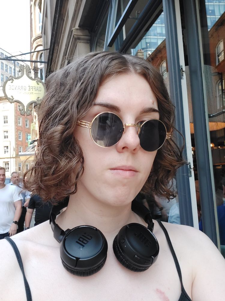 Tucker with a curly bob cut, wearing a low cut vest headphones and sunglasses