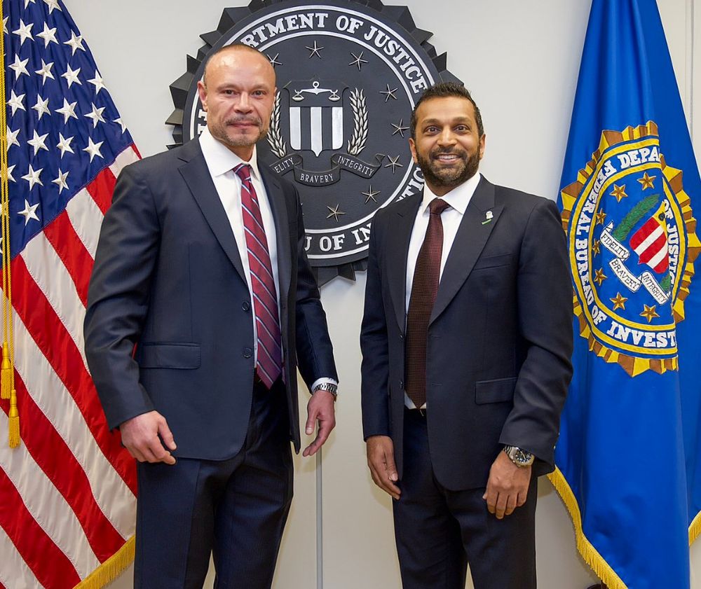 Dan Bongino (left) and Kash Patel (right) standing awkwardly in front of the Seal of the Department of Justice, an American flag, and the FBI flag.