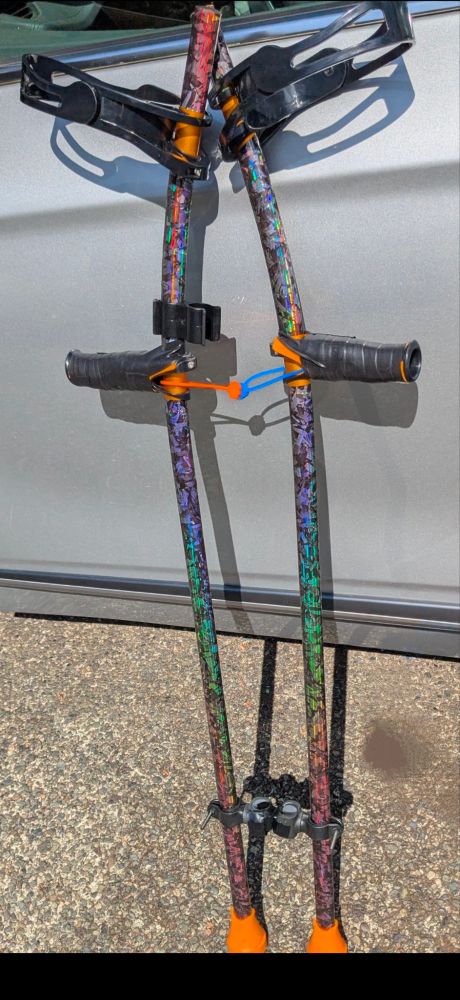 Pic of iridescent rainbow carbon fibre wrapped aluminum forearm crutches with orange ferrules and black & orange accents propped against a car door. 