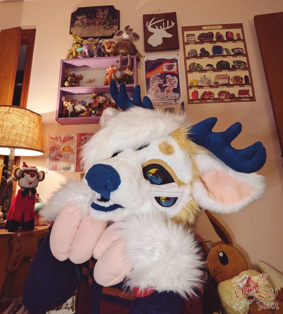 A photo of me in my fursuit, which is a sandy and white piebald whitetail deer with five teal and red eyes, pink hooves, and teal antlers and nose. I'm doing a cute post with my hooves held to my cheeks. In the background you can see some of my deer plushie collection and a giant Eevee plush.