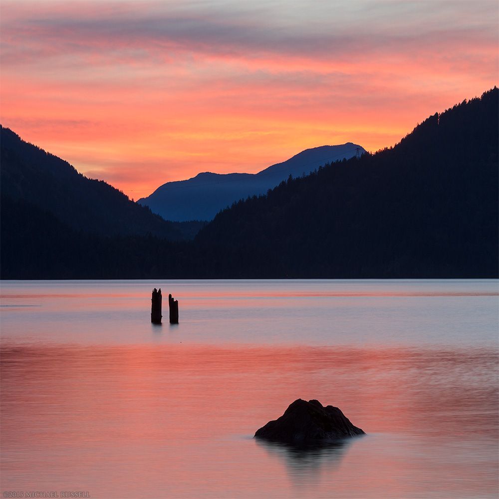 A vibrant sunset at a lake with a subtle mountain range at the other end, and two old piles and a rock in the foreground water.