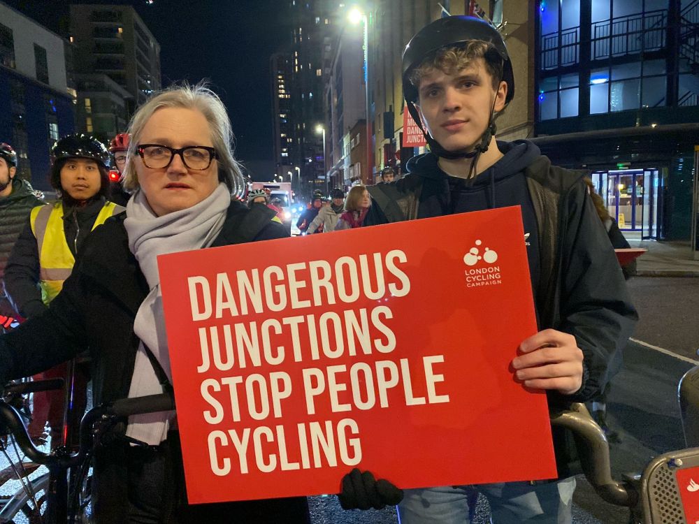 Big red sign with white text “DANGEROUS JUNCTIONS STOP PEOPLE CYCLING” me in a blue scarf and Nate in dark jacket at Lindin Cycling campaign vigil. 