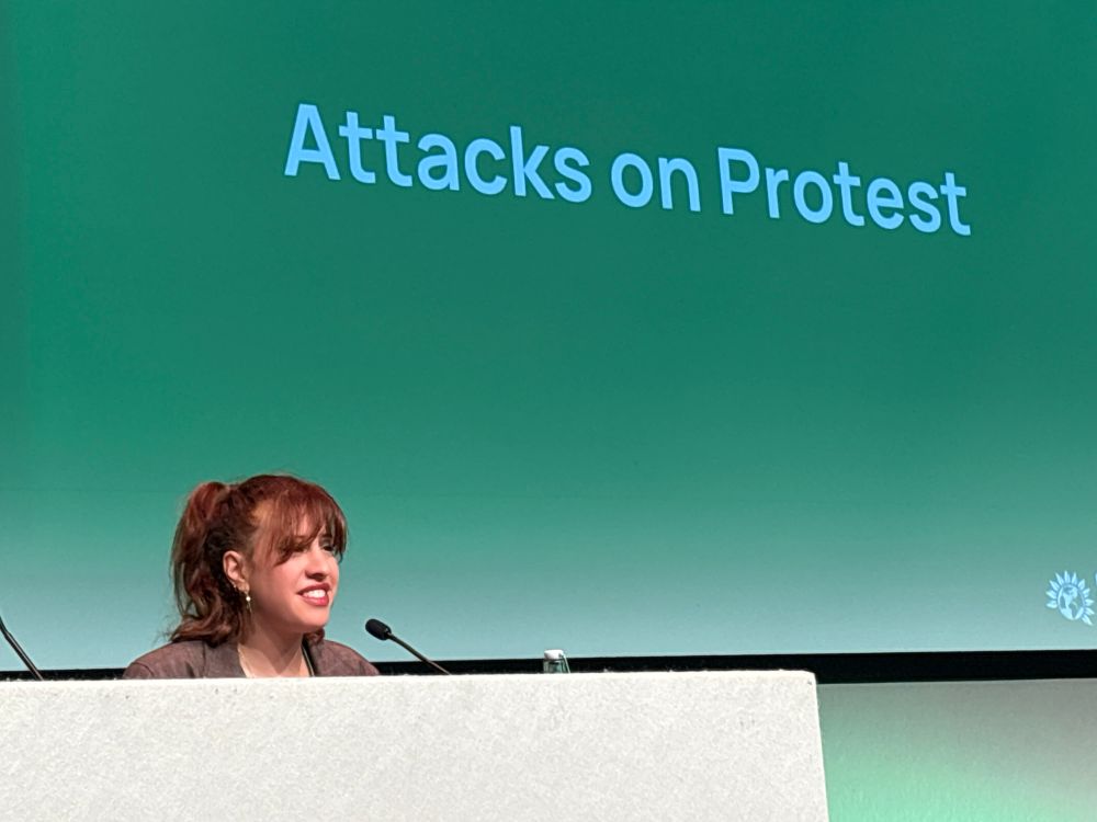 Speaker from Defend our Juries. In front of green banner saying “attacks on protest”