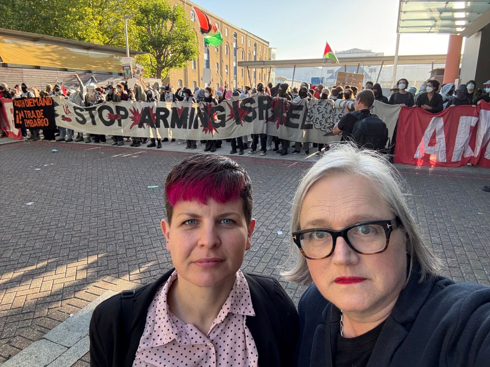 Caroline and Zoë at DSEI standing in front of a protest blocking the entrance to Excel. With big banners stop funding war crimes. Stop arming Israel. 
