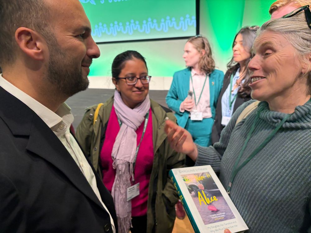 Zack and Caroline Litman smiling and speaking together, Caroline offering Zack her book - busy with people in background looking in different directions in front of Green Party branding. 