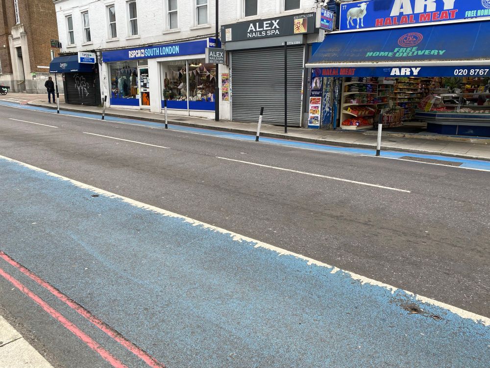 A faded blue bike lane in foreground with missing wands to protect people on bikes from traffic