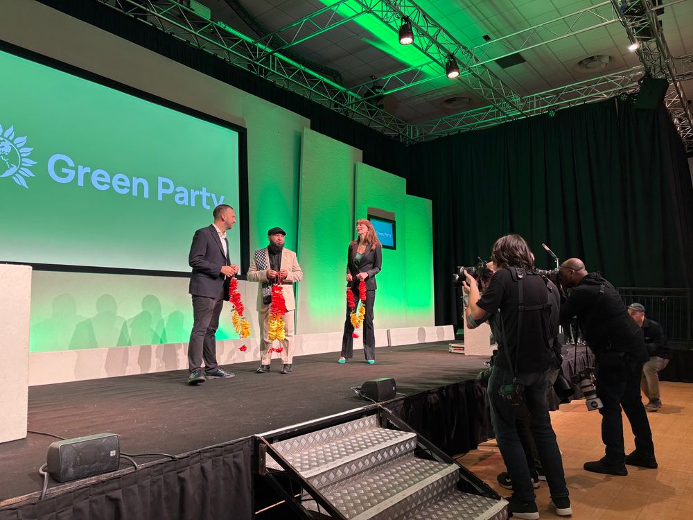 Zack, Mothin and Rachel on the conference stage, holding yellow and red garlands surrounded by photographers leaning in. Green Party branding behind them. 