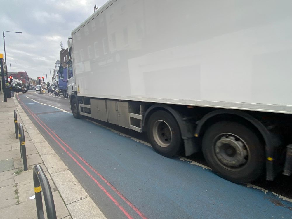 A faded blue bike lane with a huge white lorry looming across the bike lane. 
