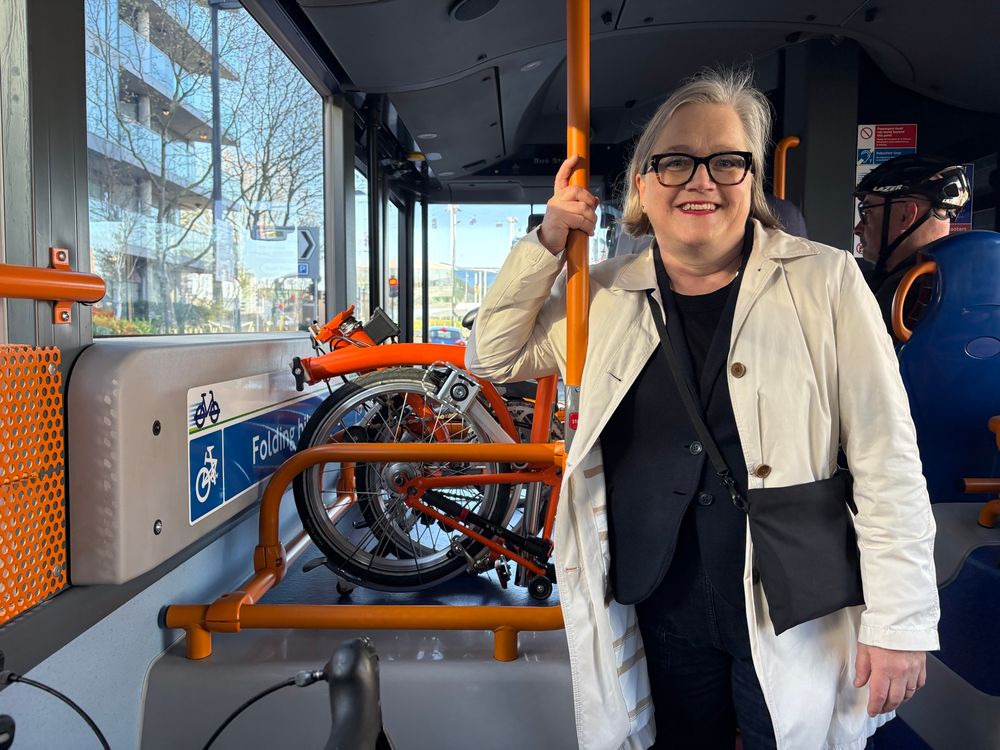 Caroline standing in white coat smiling with folded bike behind her on the bike shuttle bus. 