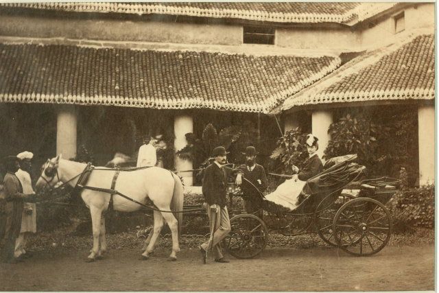 A British family in front of their Indian house, 1875.  There is a white woman in an open carriage being pulled by a white horse. There are two men in bowler hats by the carriage, on is definitely white. Two Indian men stand at the horse's head.