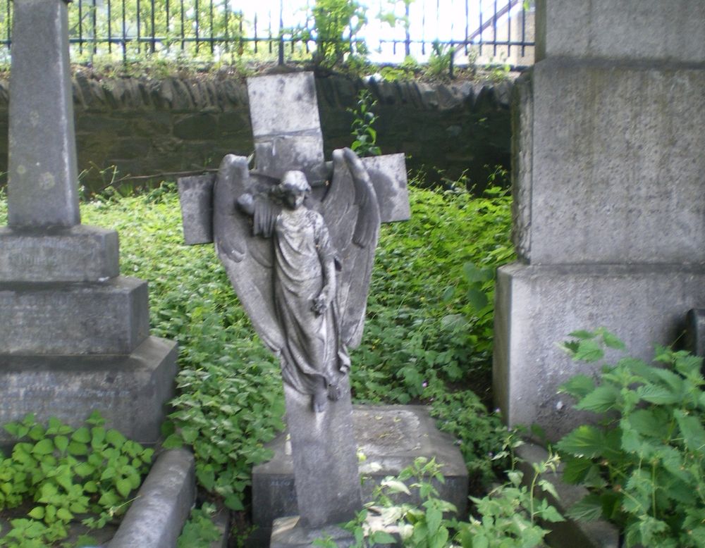 Three overgrown gravestones. The one in the middle is an angel on a lopsided cross. 
