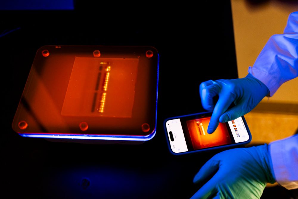 In a dark lab room, two screens light up red on a desk, with blue gloved hands touching the phone screen.