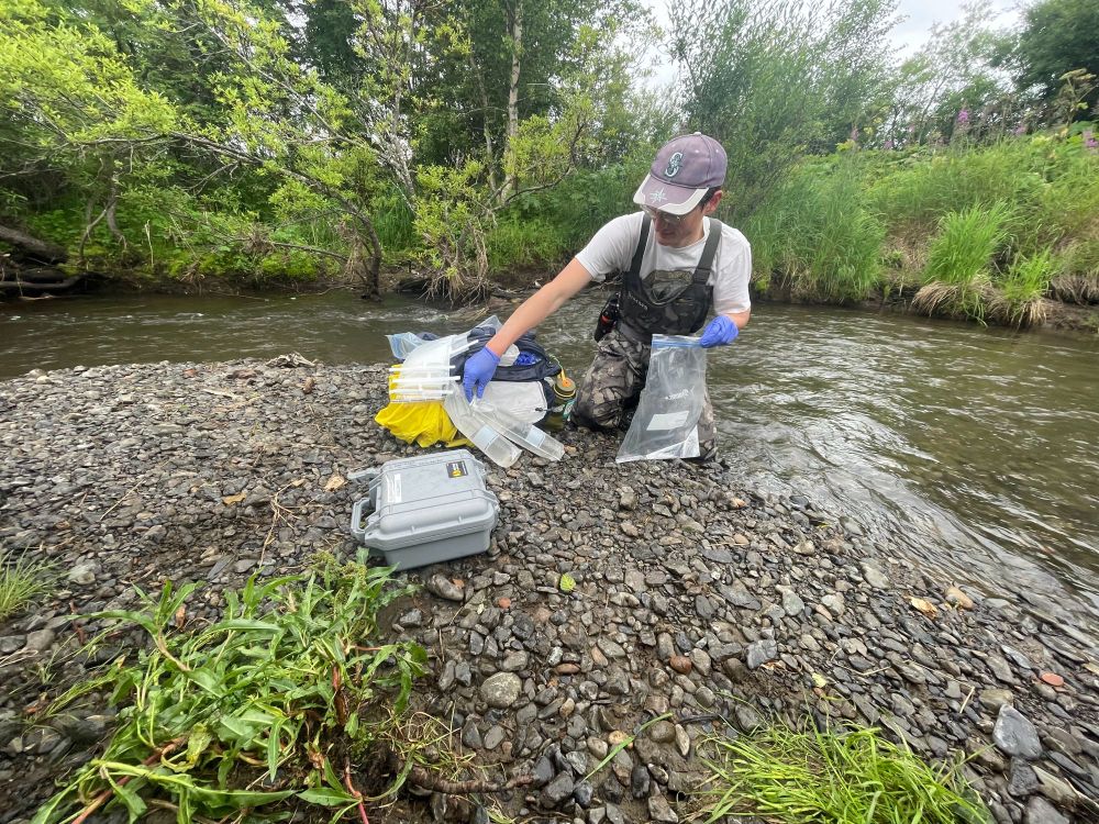 Ryan kneels on the side of a stream in Alaska, with equipment to collect eDNA samples.