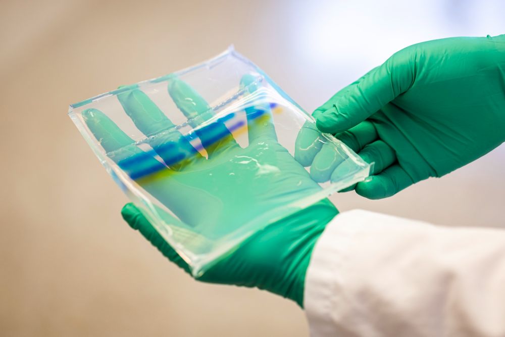 Hands wearing green gloves hold a square clear dish with ribbons of color.