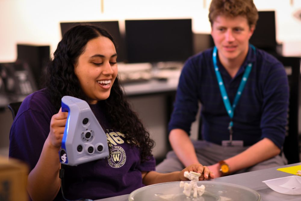 Eliana Shankar (l) and Callum Backstrom (r) sit at a table. Eliana holds a scanner over a piece of coral