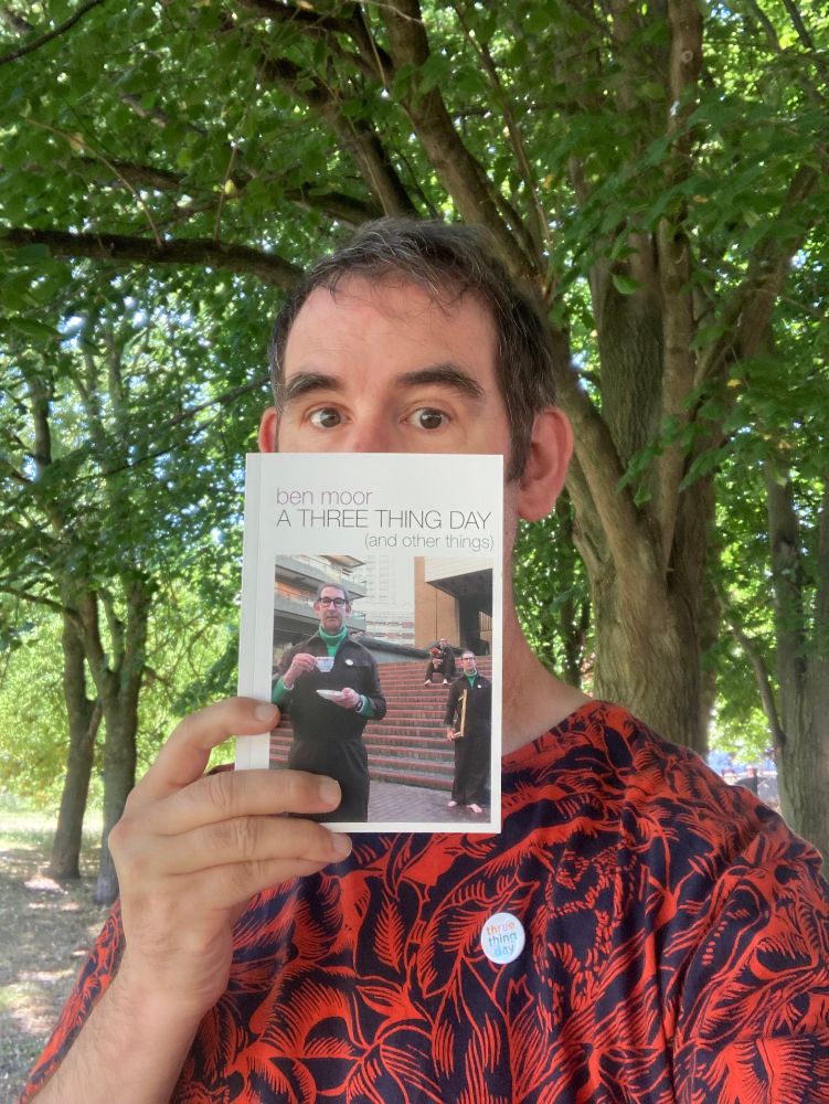 Actor/Writer Ben Moor holding a copy of his book A Three Thing Day (and other things) among some trees.
