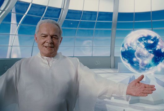 Othon Bastos com uma túnica branca recebendo uma pessoa de braços abertos. Ao fundo, um cenário meio futurista azul com janelas e um globo terrestre