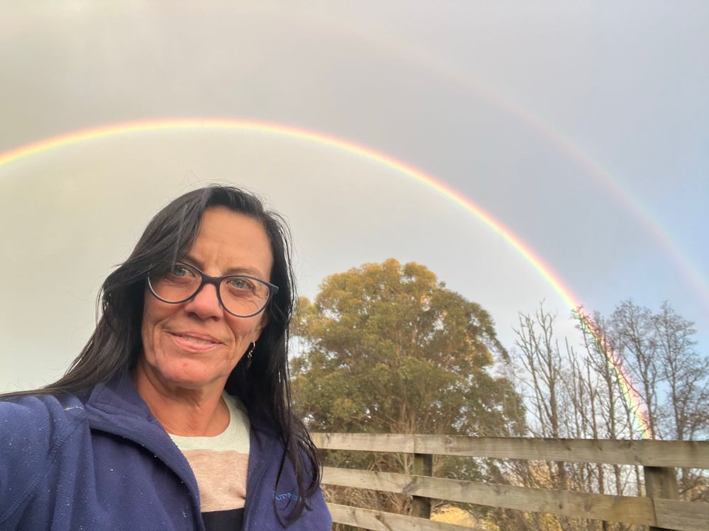 Dark haired woman with glasses in the foreground and a double rainbow in the sky behind 