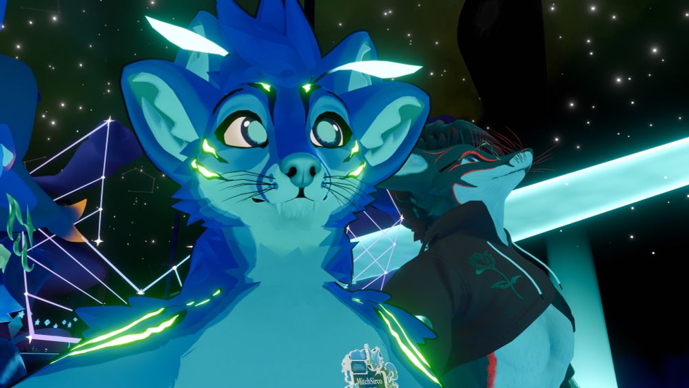 A blue four eared Fennix (me) looking at a screen outside of this image. On the right, an additional person is seen (its DrBlackRat). The currently playing set is from loprov